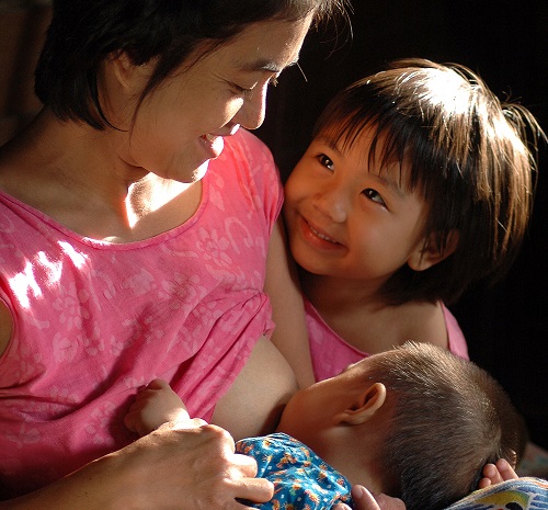 woman breastfeeding a baby with an older child looking on